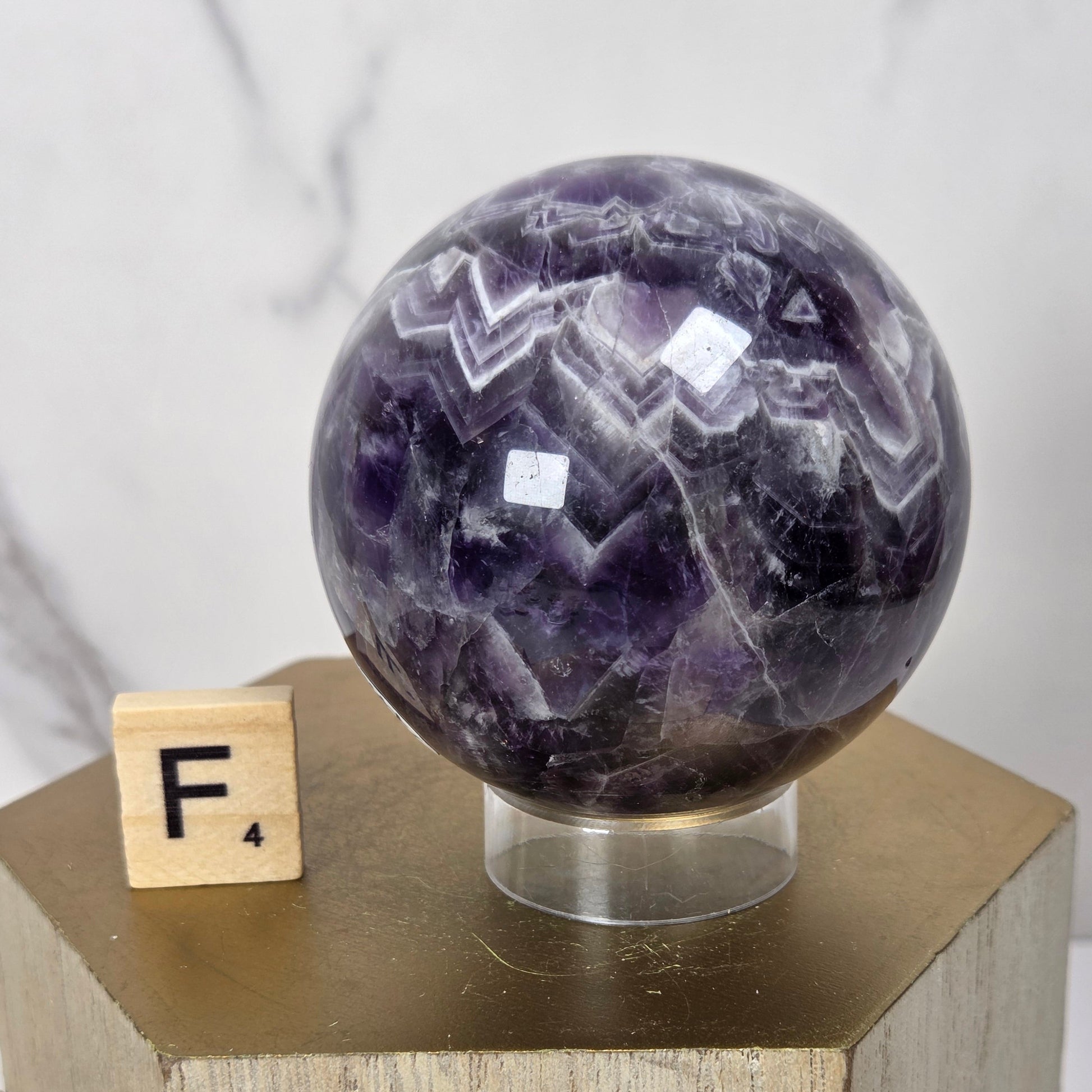A polished purple amethyst crystal sphere displayed on a clear acrylic stand atop a light wood block, with a small wooden letter tile showing 'F' beside it against a light marble-patterned background.