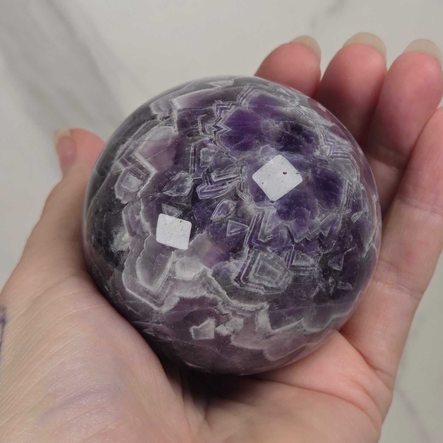A hand holding a polished purple amethyst crystal sphere with white geometric inclusions, set against a light marble background.