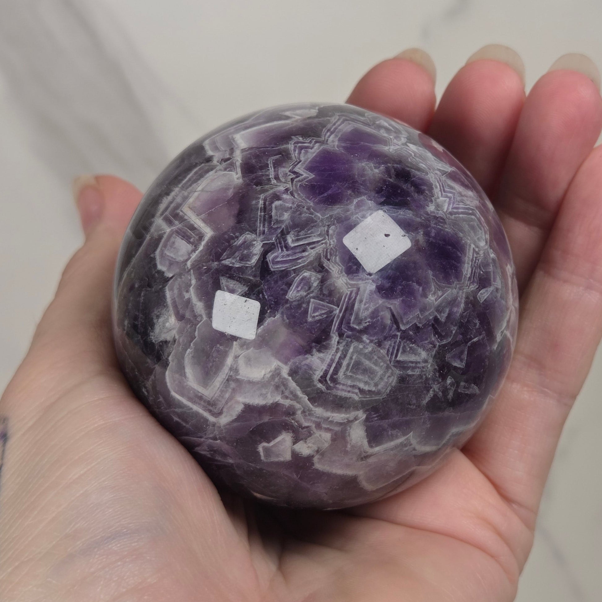 A hand holding a polished purple amethyst crystal sphere with white geometric inclusions, set against a light marble background.