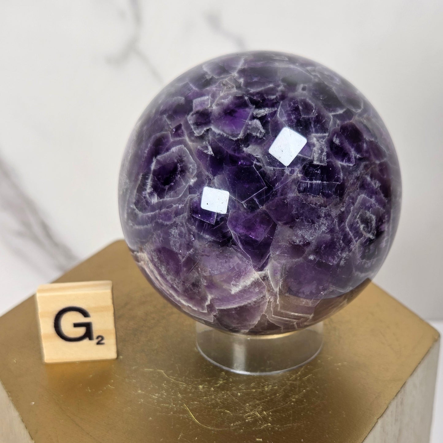 A polished purple amethyst crystal sphere displayed on a small clear stand atop a gold hexagonal pedestal, with a wooden Scrabble tile showing 'G2' beside it.