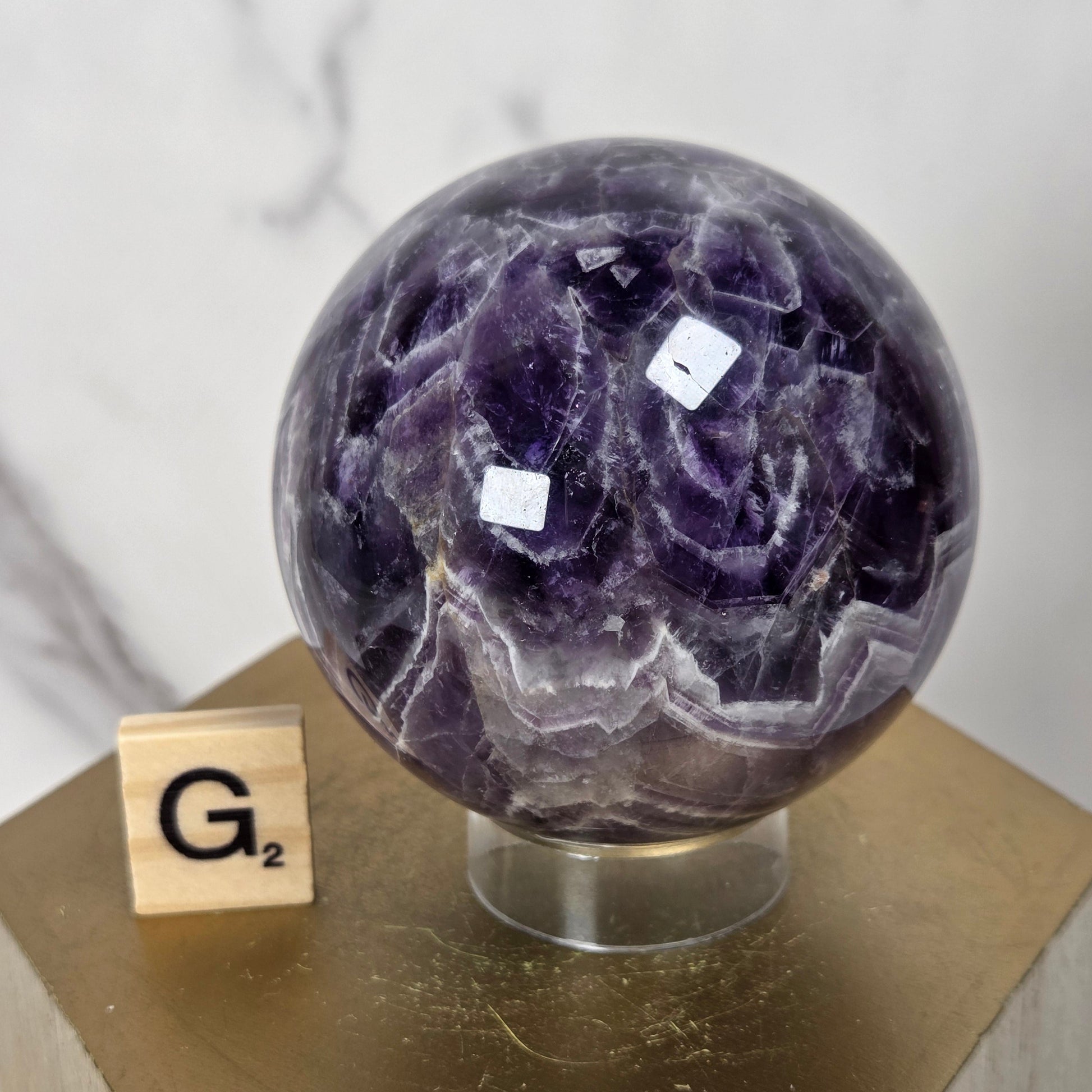 A polished purple amethyst geode sphere on a clear acrylic stand, displayed on a gold-colored wooden pedestal next to a small wooden Scrabble tile showing 'G2'.