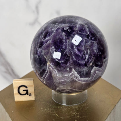 A polished purple amethyst geode sphere on a clear acrylic stand, displayed on a gold-colored wooden pedestal next to a small wooden Scrabble tile showing 'G2'.