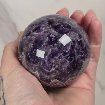 A hand holds a polished purple amethyst crystal sphere with visible natural crystalline texture and two small white stickers on the surface, against a light marble-patterned background.