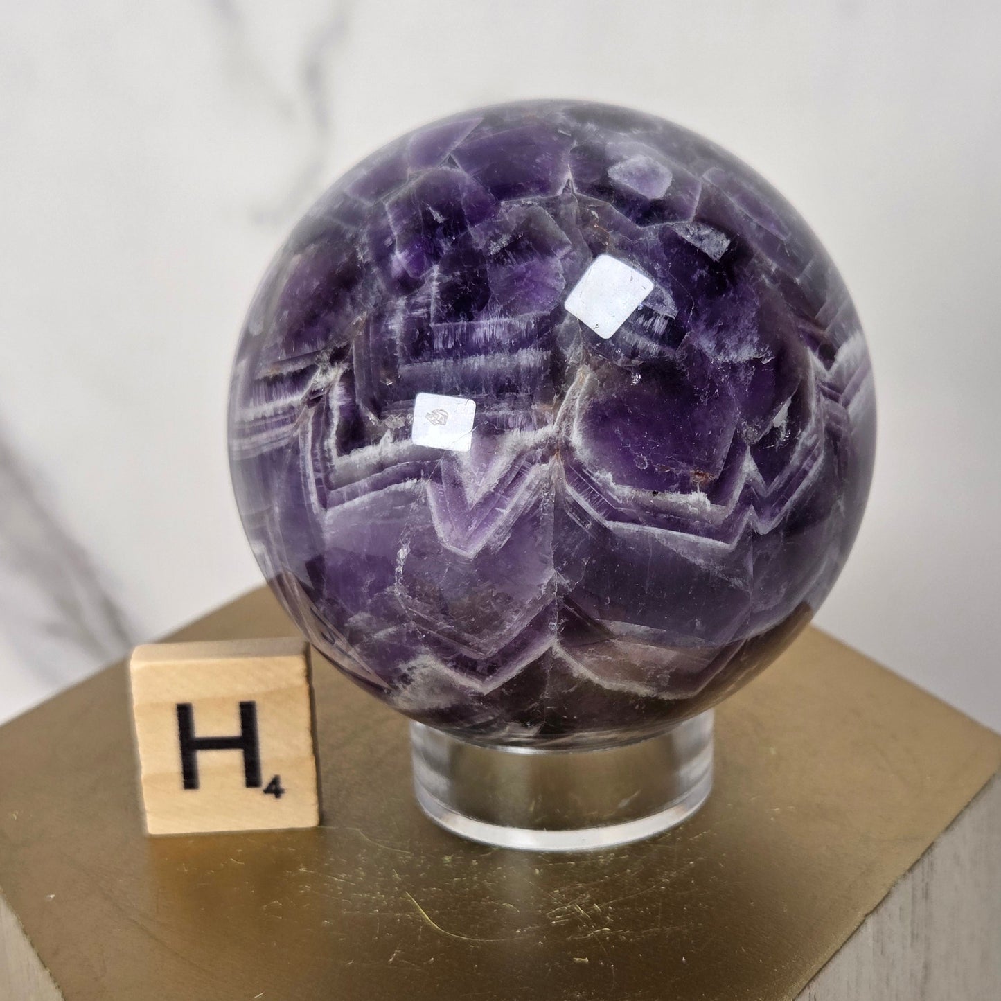 A polished purple amethyst geode sphere displayed on a clear acrylic stand atop a gold hexagonal block, with a small wooden Scrabble tile showing the letter H in the background against a light marble wall.