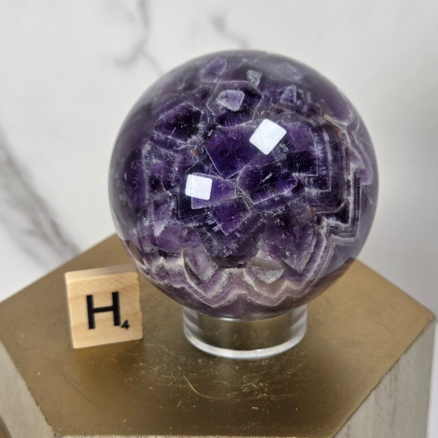 A polished purple amethyst crystal sphere displayed on a clear glass stand atop a gold hexagonal pedestal, with a small wooden letter tile showing 'H' beside it against a light marble-patterned background.