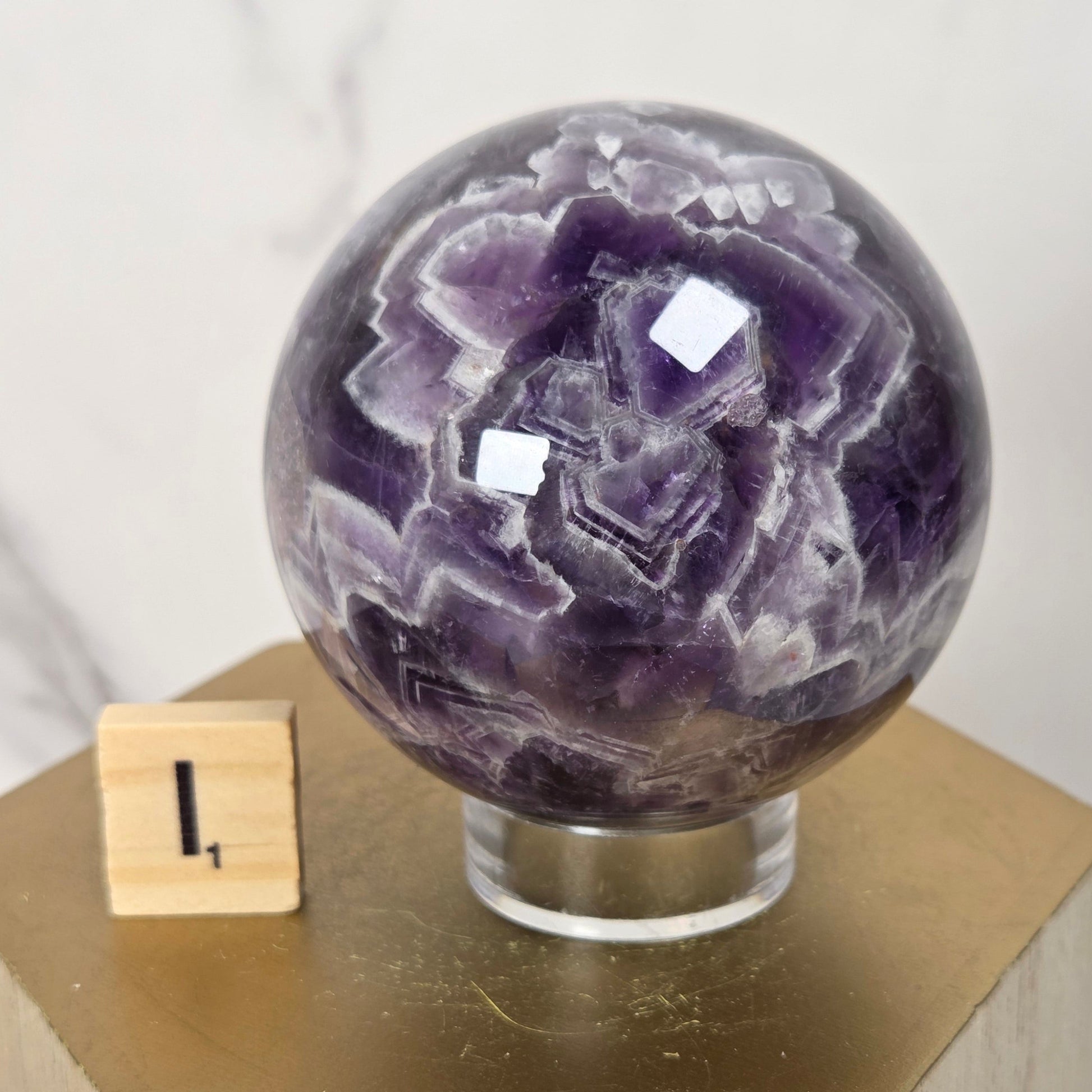 A polished purple amethyst crystal sphere on a clear acrylic stand, displayed on a gold hexagonal pedestal with a small wooden block showing the number one.