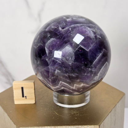 A polished purple amethyst crystal sphere displayed on a clear acrylic stand atop a gold hexagonal pedestal, with a small wooden block showing the number one in the background.