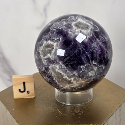 A polished purple amethyst crystal sphere with white and gray marbling, displayed on a clear stand atop a gold hexagonal pedestal. A small wooden Scrabble tile with the letter J sits beside it against a light marble-patterned background.