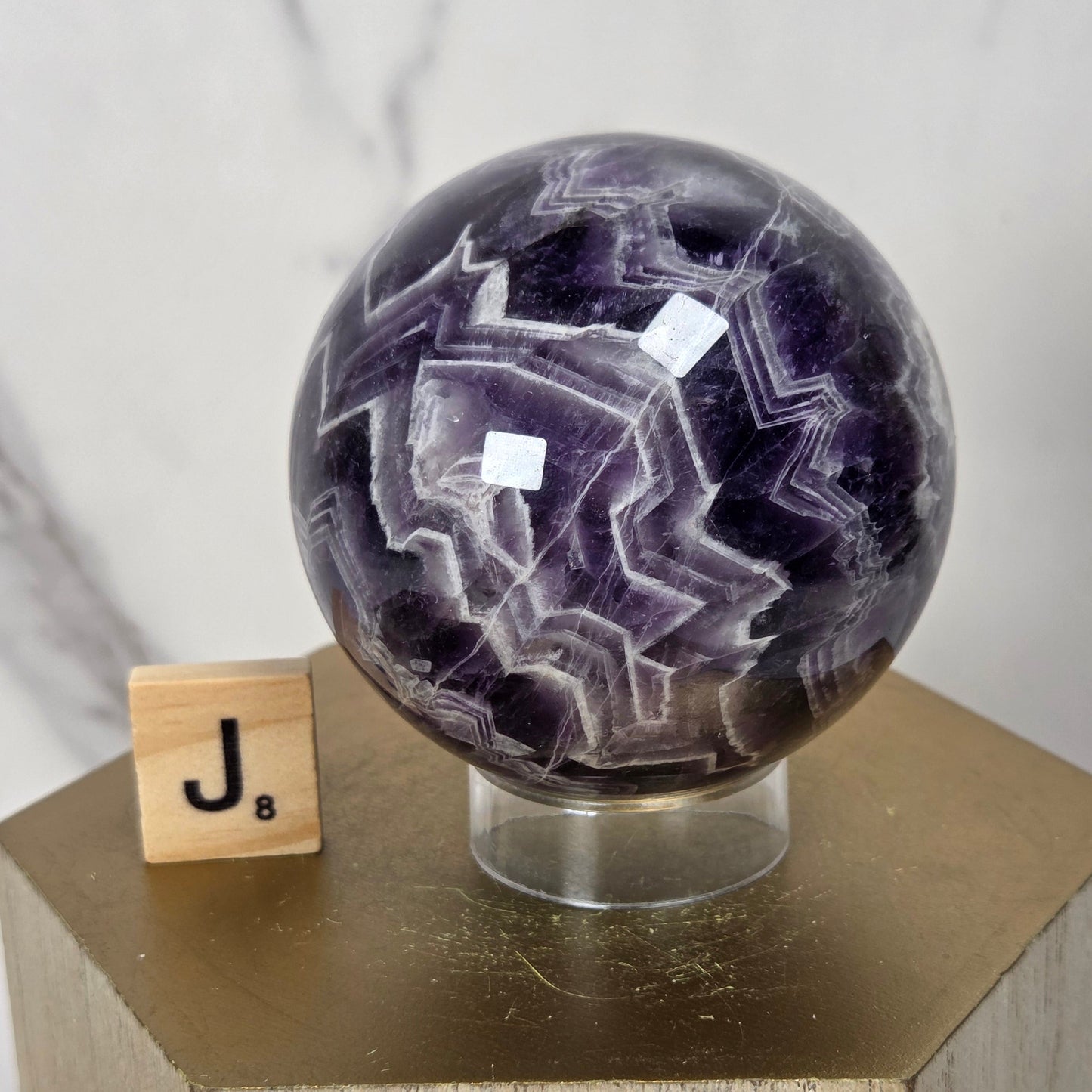 A polished purple and white agate crystal sphere on a clear acrylic stand, displayed on a hexagonal wooden pedestal next to a wooden letter tile with the letter J.