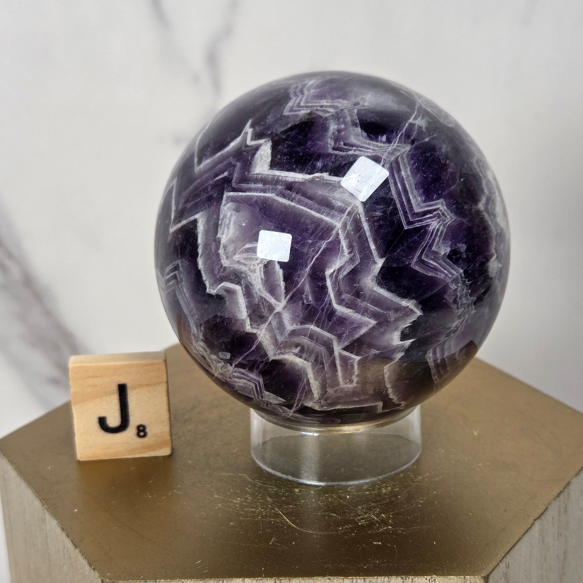 A polished purple and white agate crystal sphere on a clear acrylic stand, displayed on a hexagonal wooden pedestal next to a wooden letter tile with the letter J.