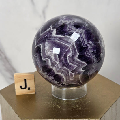 A polished purple and white agate crystal sphere on a clear acrylic stand, displayed on a hexagonal wooden pedestal next to a wooden letter tile with the letter J.