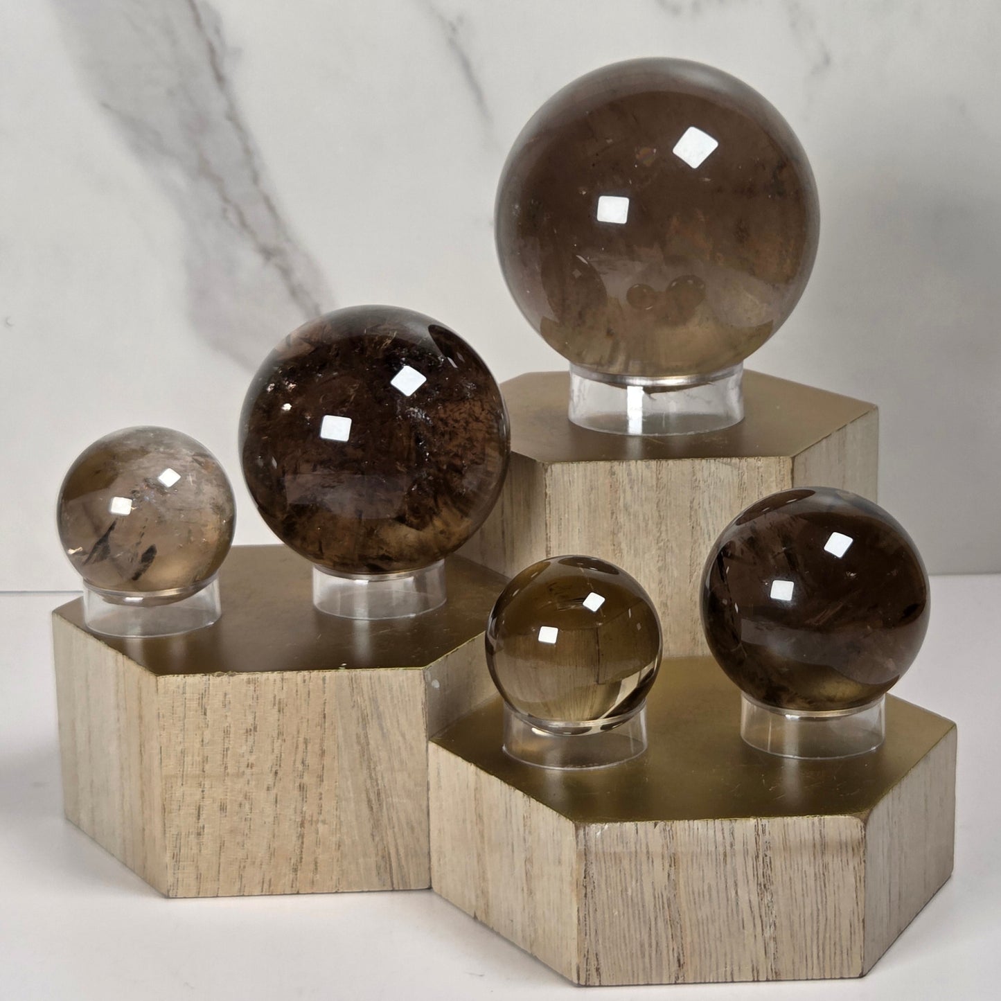 Five polished crystal spheres in varying sizes and smoky brown tones, each mounted on a clear stand and displayed on a light wood hexagonal riser against a white marble background.