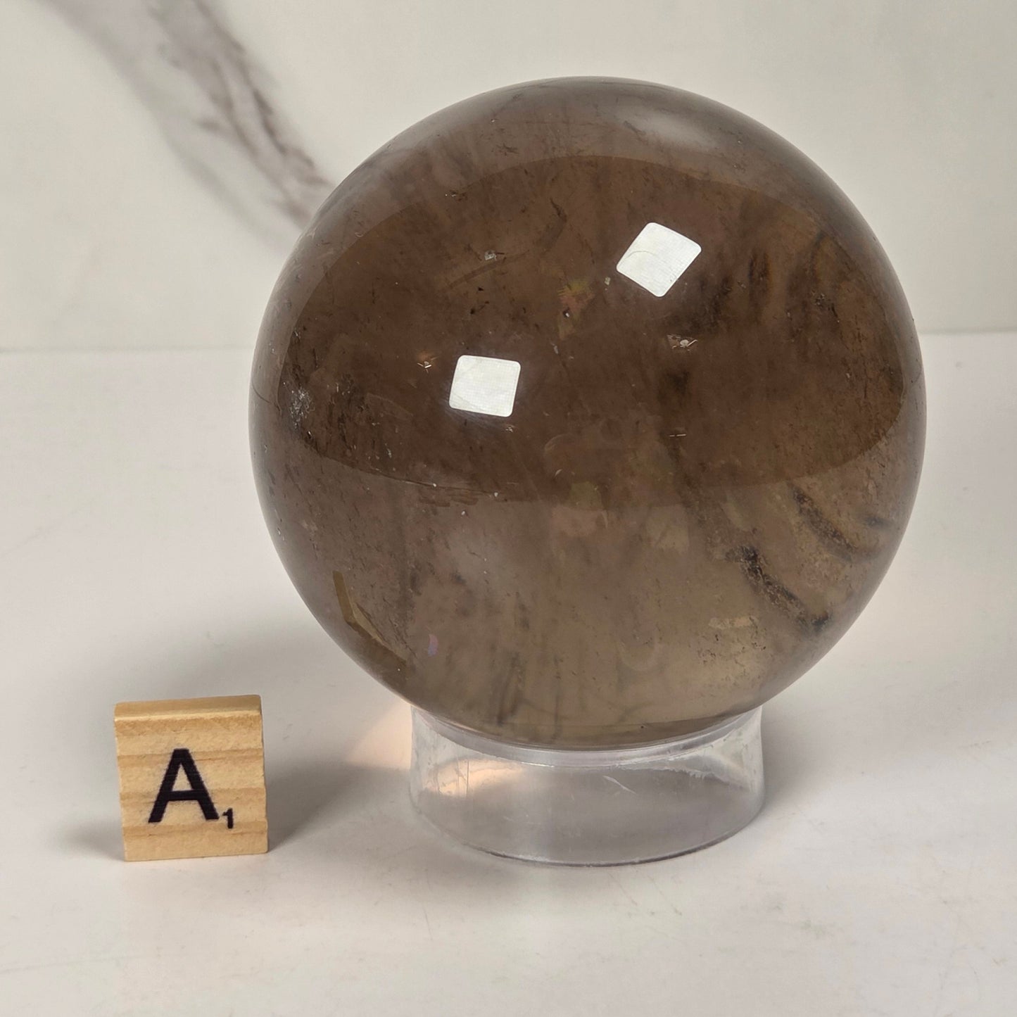 A polished brown smoky quartz crystal sphere on a clear acrylic stand, displayed next to a small wooden Scrabble tile on a white marble surface.