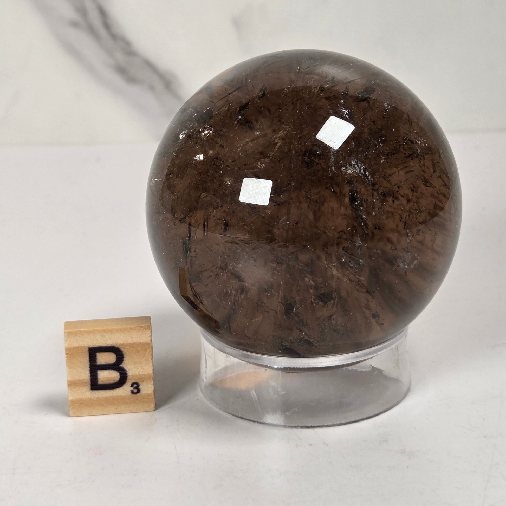 A polished dark brown crystal sphere on a clear acrylic stand, shown next to a wooden Scrabble tile spelling B³ on a light marble surface.