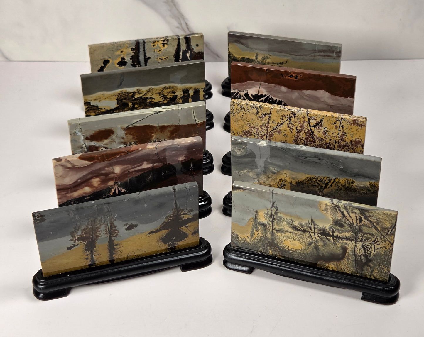 A set of 12 small landscape art slabs, each mounted on a black display stand, arranged in two columns against a white marble-patterned background.