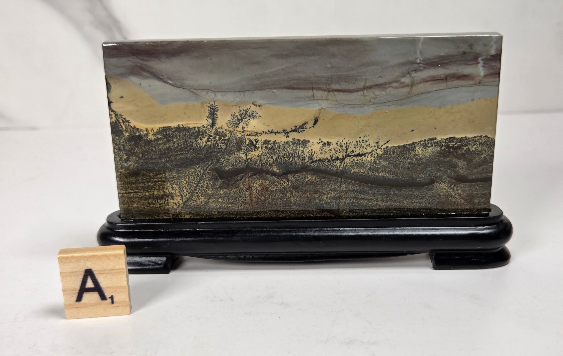 A rectangular stone or wood plaque with a landscape-like marbled pattern in gray, beige, and brown tones, displayed on a black stand. A small wooden letter tile with the letter A is placed in front on a white surface.