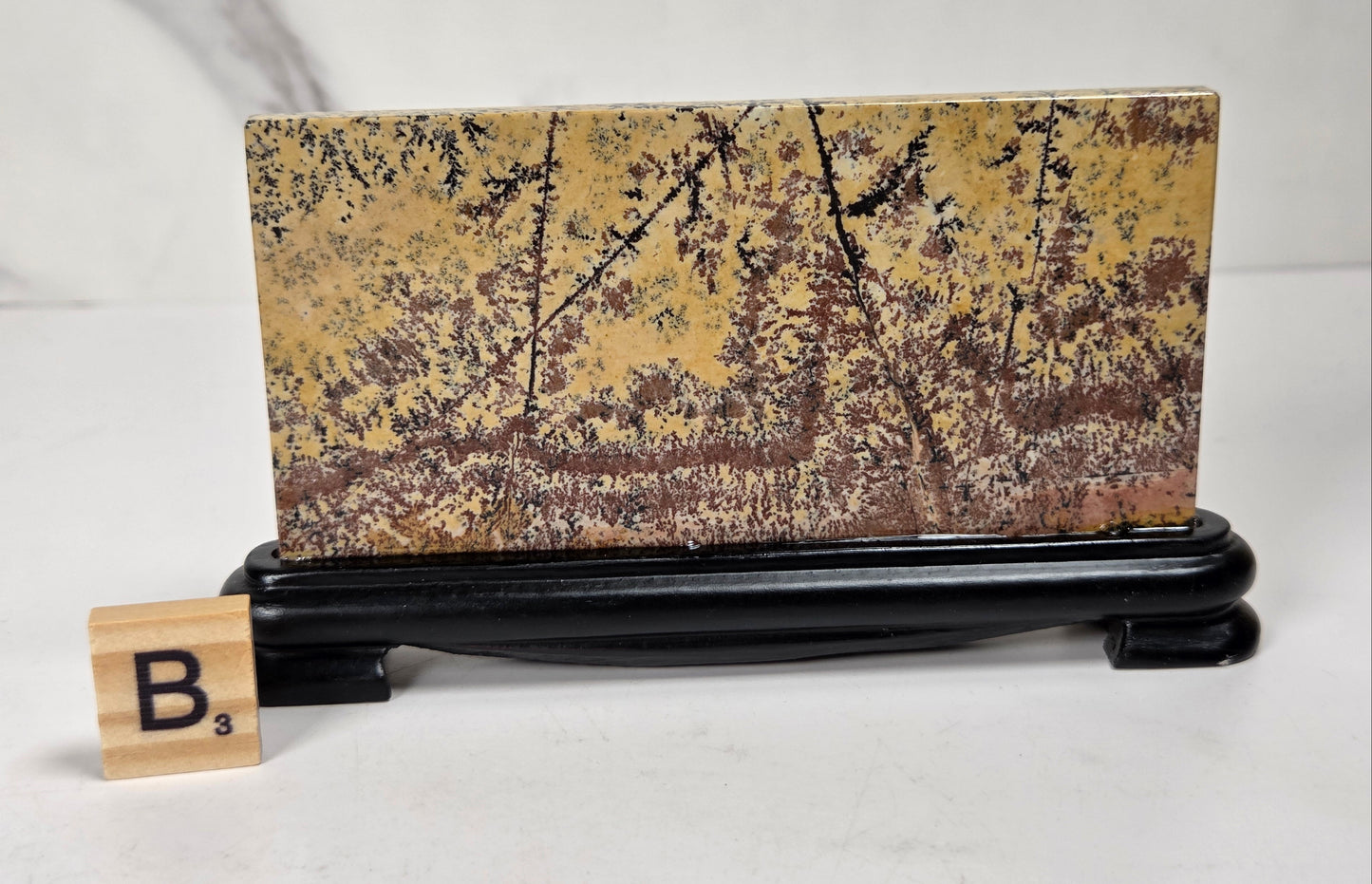A rectangular decorative plaque or tile with a mottled gold, brown, and black abstract pattern, displayed upright on a black stand. A small Scrabble tile with the letter B is placed in front for size reference, set against a light marble-patterned background.