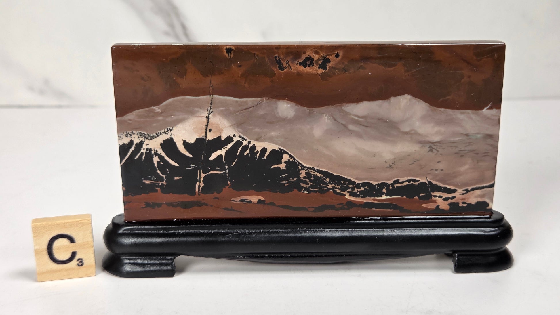 A rectangular stone or wood plaque featuring a mountain landscape scene in earthy brown, beige, black, and white tones, displayed on a black stand against a light marble-like background. A small wooden letter tile with the letter C is visible to the left.