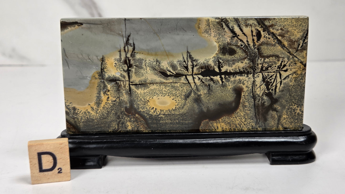 A rectangular stone or wood block with a marbled landscape pattern in gray, gold, and brown, displayed on a black stand with a small wooden letter tile showing 'D2' in front, against a light gray background.