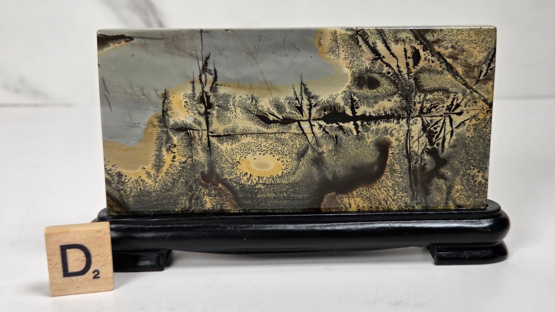 A rectangular stone or wood block with a marbled landscape pattern in gray, gold, and brown, displayed on a black stand with a small wooden letter tile showing 'D2' in front, against a light gray background.