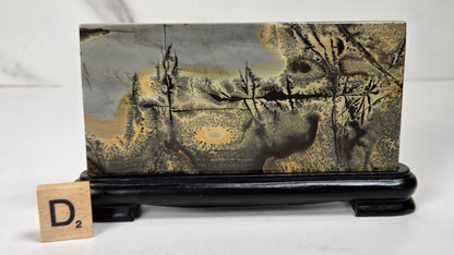 A rectangular stone or wood block with a marbled landscape pattern in gray, gold, and brown, displayed on a black stand with a small wooden letter tile showing 'D2' in front, against a light gray background.
