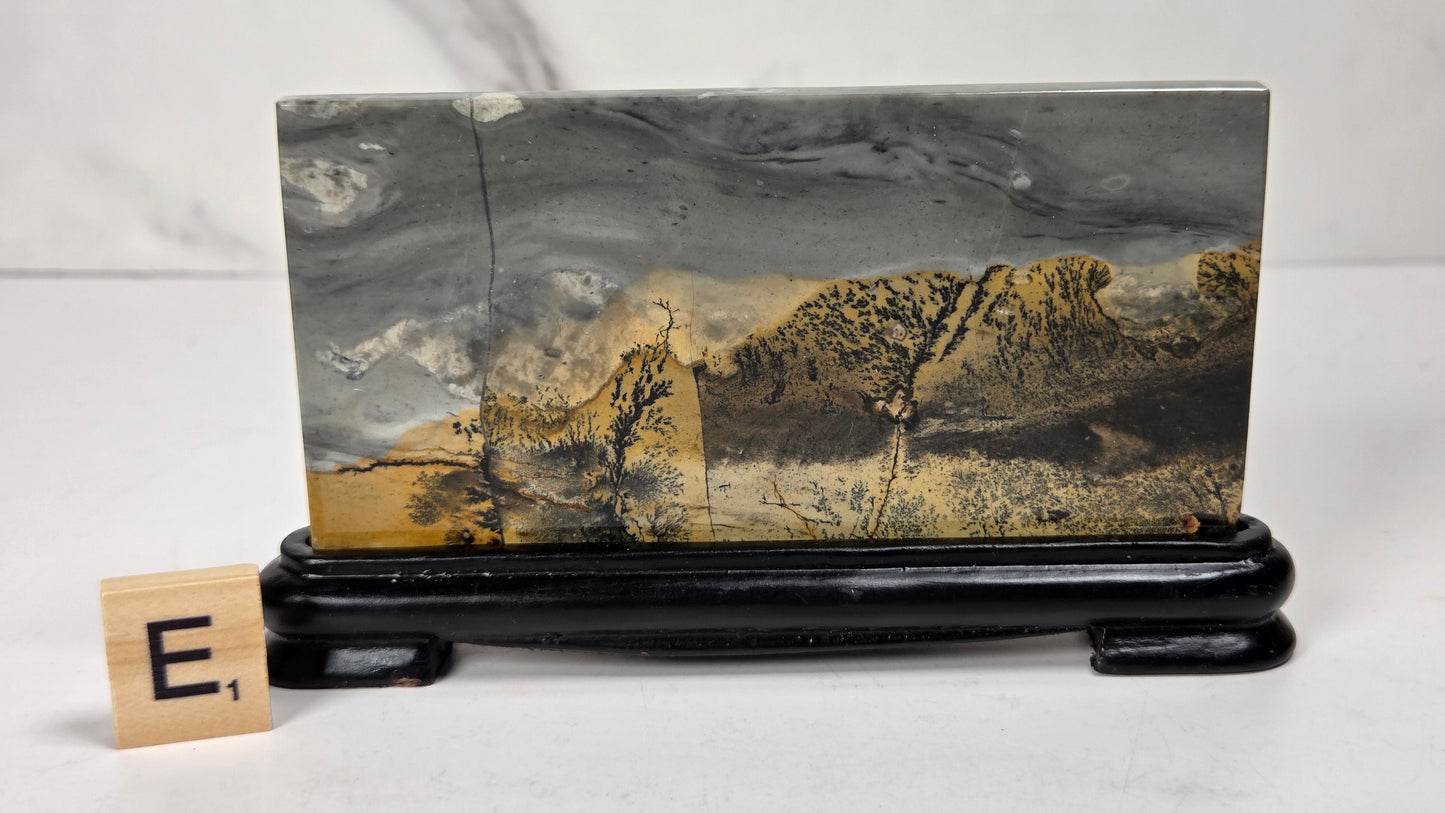 A rectangular stone or wood plaque featuring a mountain landscape print in gray, black, and golden brown tones, displayed on a glossy black stand with a small letter tile showing 'E' beside it.