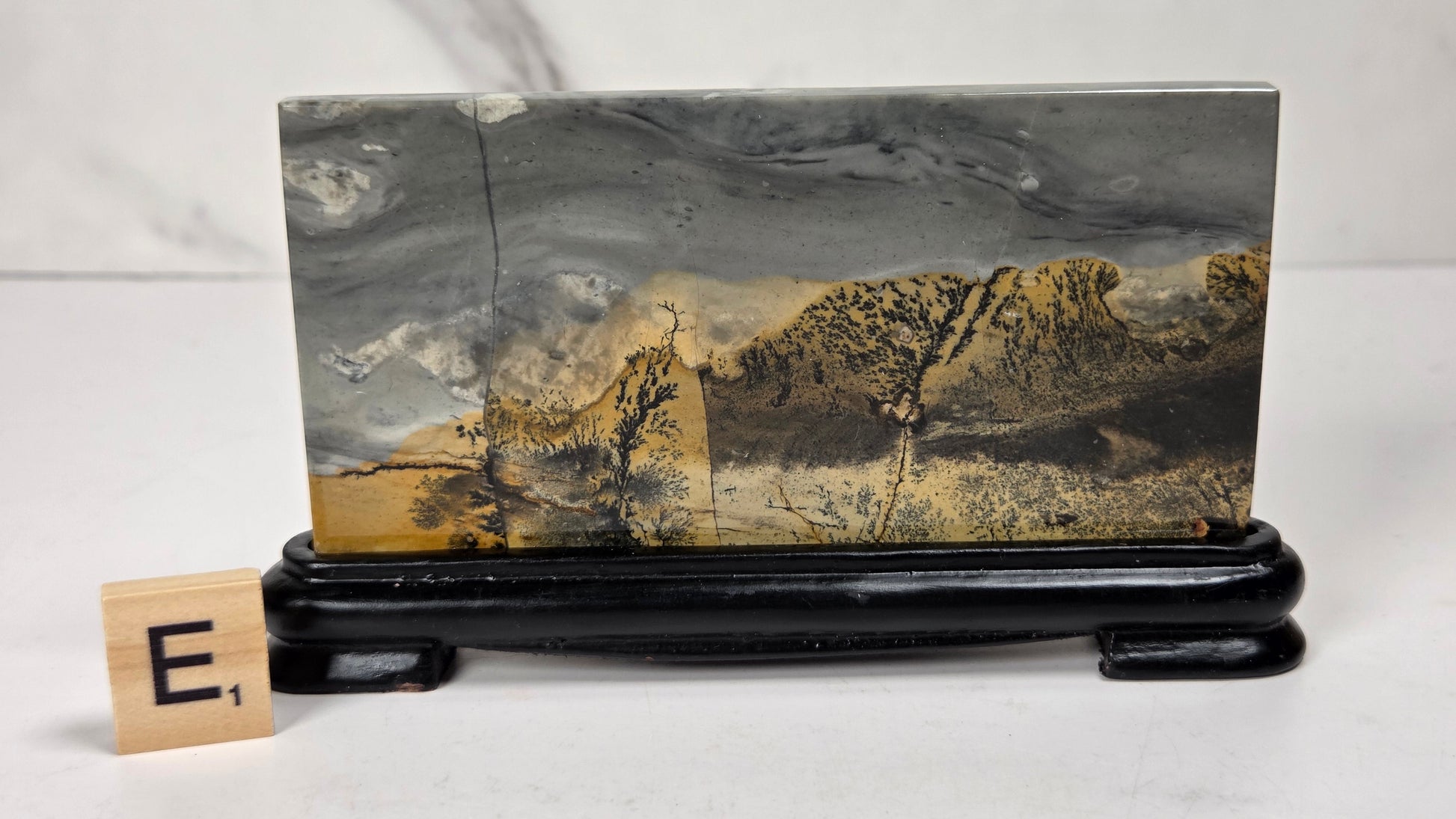 A rectangular stone or wood plaque featuring a mountain landscape print in gray, black, and golden brown tones, displayed on a glossy black stand with a small letter tile showing 'E' beside it.