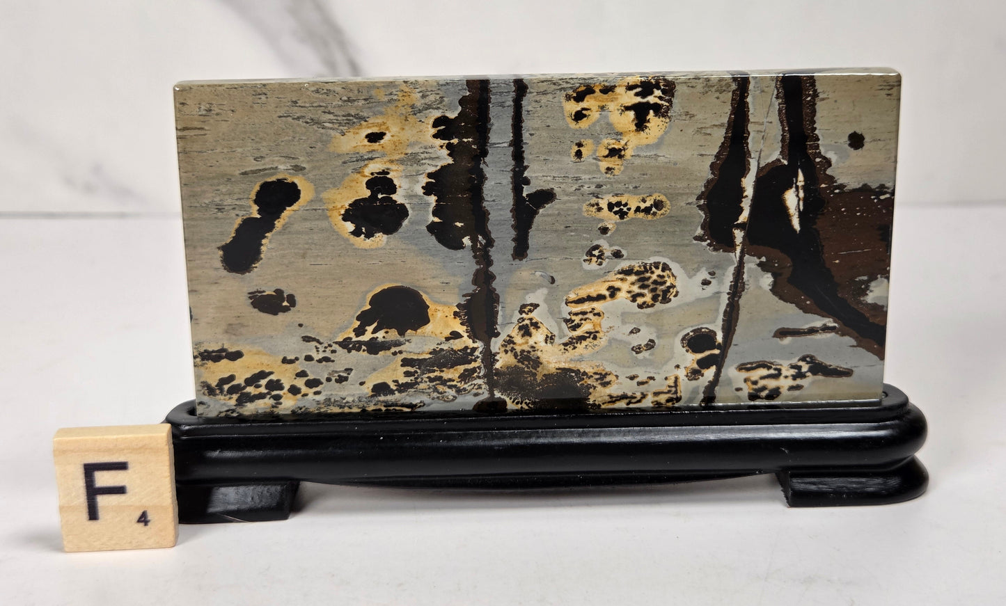 A rectangular ceramic tile with an abstract black, gold, and gray marbled pattern, displayed on a black wooden stand next to a Scrabble tile showing the letter F.