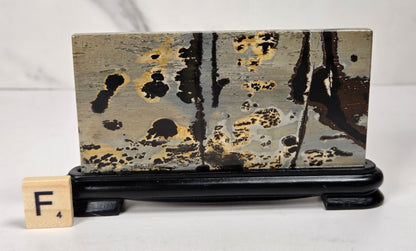 A rectangular ceramic tile with an abstract black, gold, and gray marbled pattern, displayed on a black wooden stand next to a Scrabble tile showing the letter F.