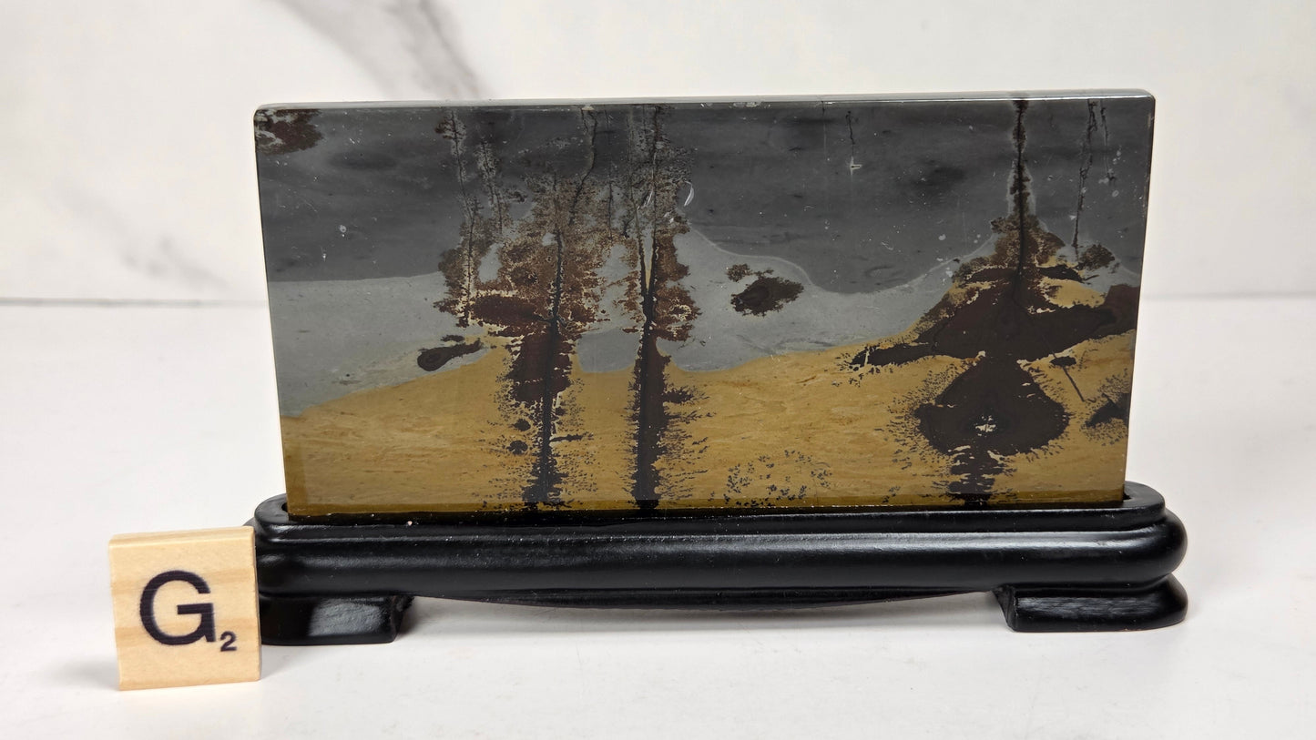 A rectangular decorative plaque or art piece with an abstract landscape design in black, gray, and gold, displayed on a black stand. A small wooden Scrabble tile with the letter G2 sits to the left for scale, set against a light gray marble-patterned background.