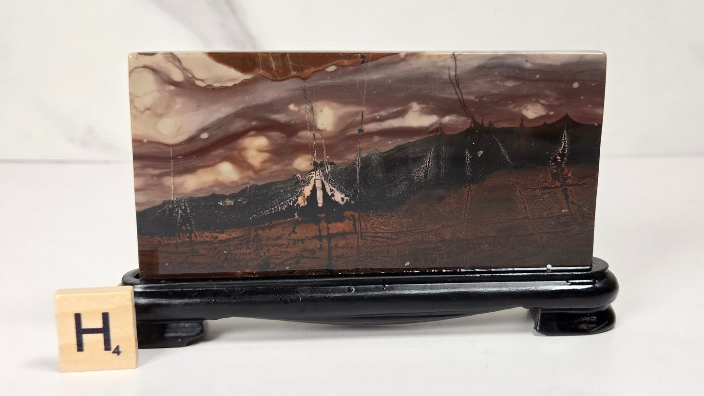 A rectangular polished agate stone slab with a mountain landscape pattern in earthy brown, beige, and black tones, displayed on a black stand with a small Scrabble tile showing 'H4' in front, set against a light gray marble background.
