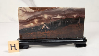 A rectangular polished agate stone slab with a mountain landscape pattern in earthy brown, beige, and black tones, displayed on a black stand with a small Scrabble tile showing 'H4' in front, set against a light gray marble background.