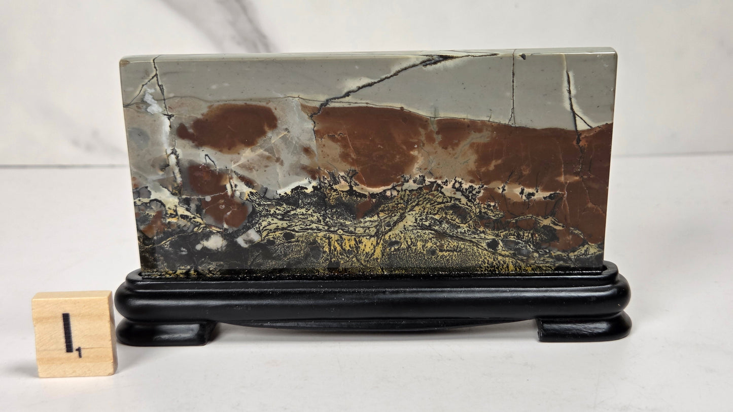 A rectangular polished stone slab with a landscape-like marbled pattern in brown, gray, black, and gold, displayed on a black stand next to a small wooden block with the number 1.