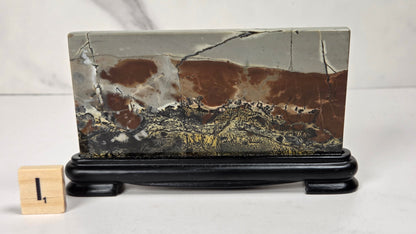 A rectangular polished stone slab with a landscape-like marbled pattern in brown, gray, black, and gold, displayed on a black stand next to a small wooden block with the number 1.