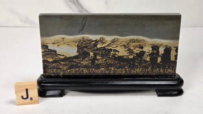 A rectangular landscape art plaque featuring a scenic mountain and lake landscape in black, gold, and gray tones, displayed on a black stand with a small wooden letter block showing 'J8' on a light marble surface.