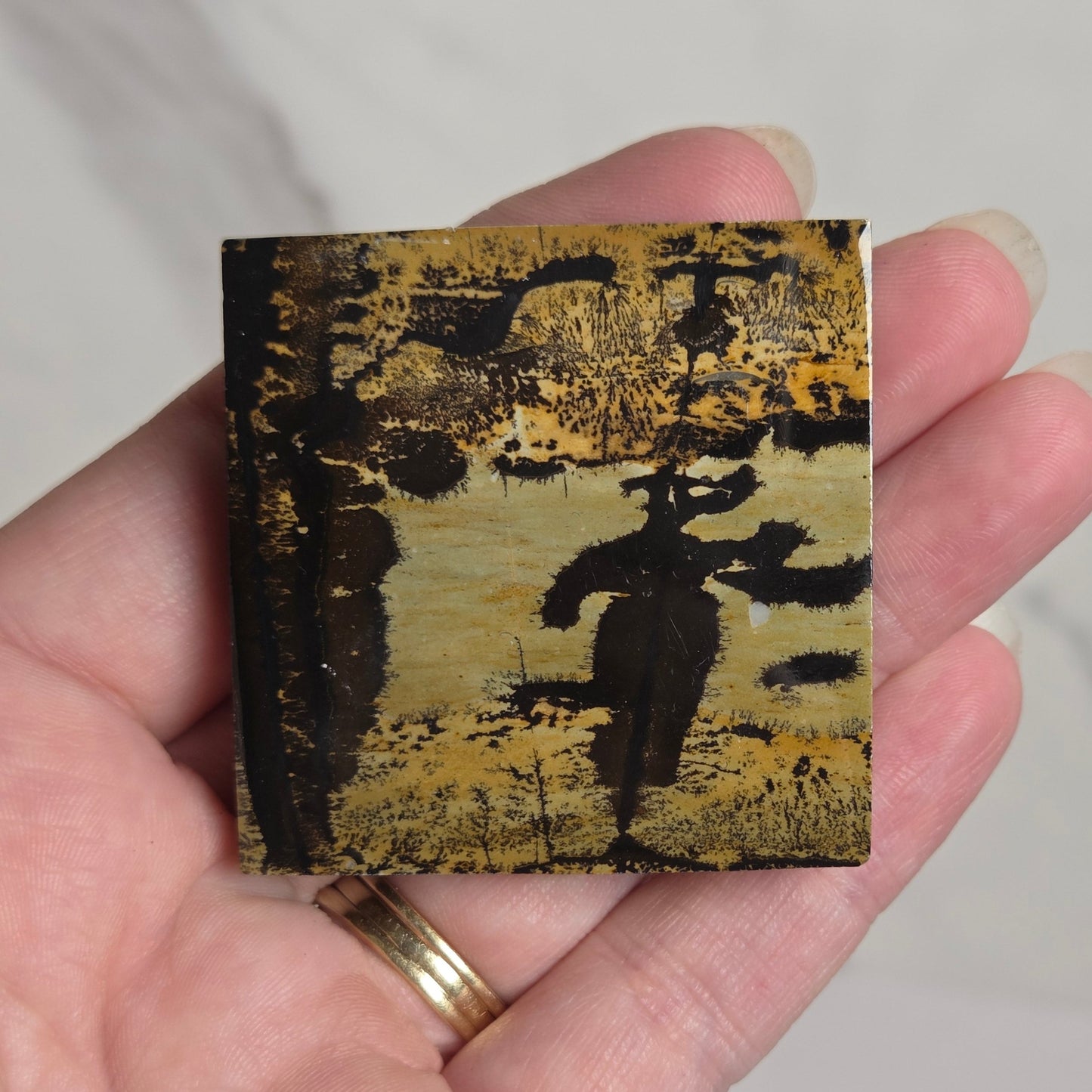 A hand holding a small square tile featuring a textured, abstract landscape painting in black and gold tones, set against a light marble background.
