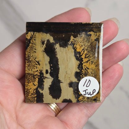 A hand holding a small square ceramic tile with an abstract black and gold marbled pattern, featuring a white circular sticker that reads '10 Jul'.