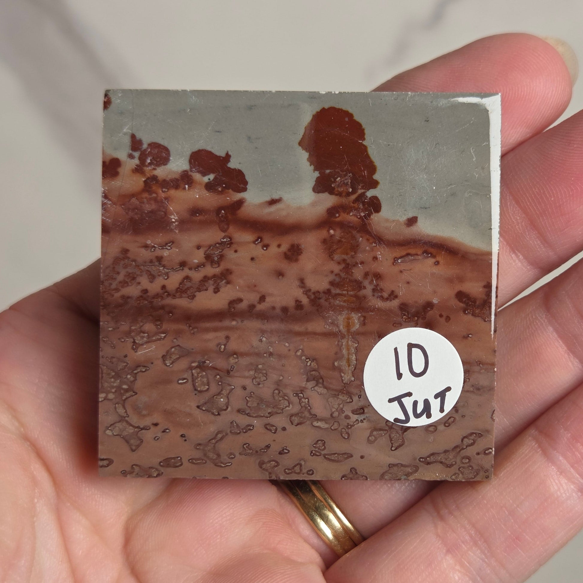 A hand holding a square ceramic tile or coaster showing a mottled brown and gray pattern, with a white circular sticker reading '10 JUT'.