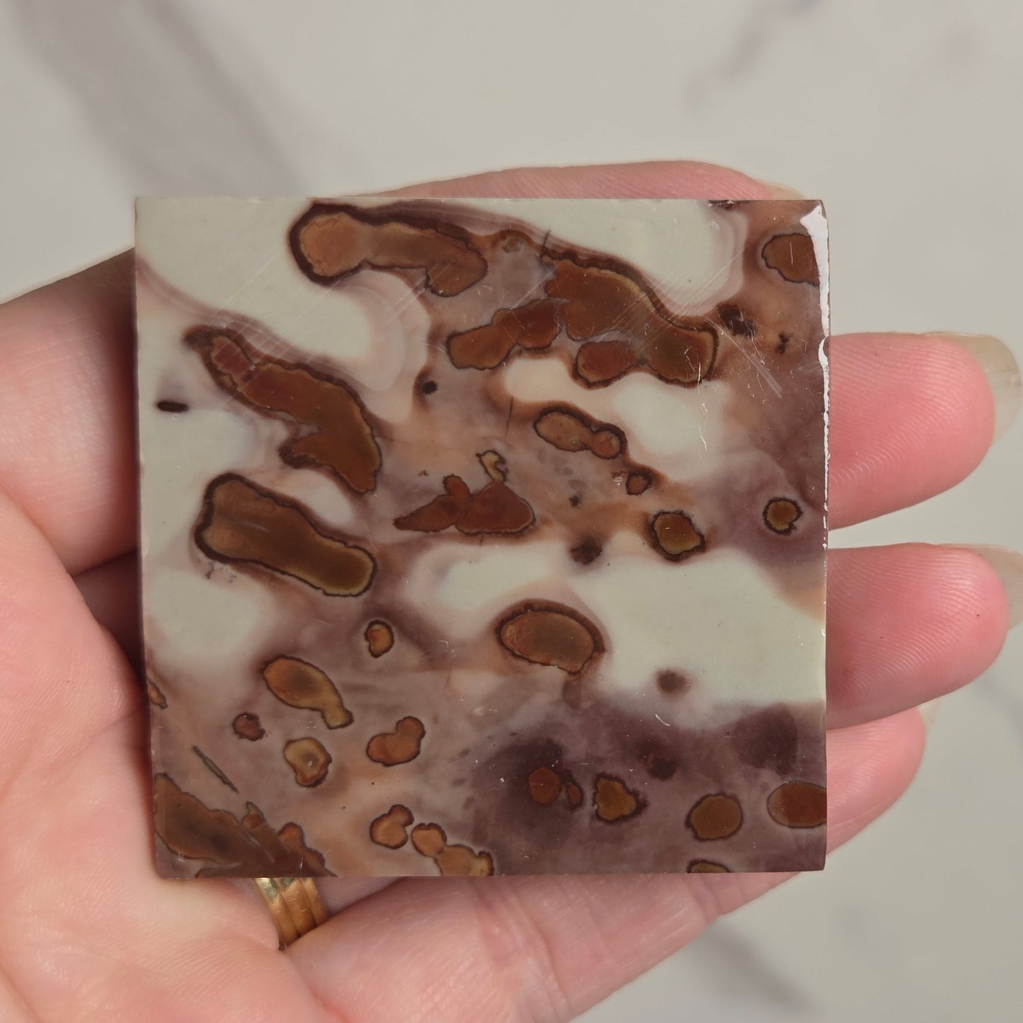 A hand holding a square decorative tile or coaster with a marbled pattern of brown, beige, and white, set against a light marble background.