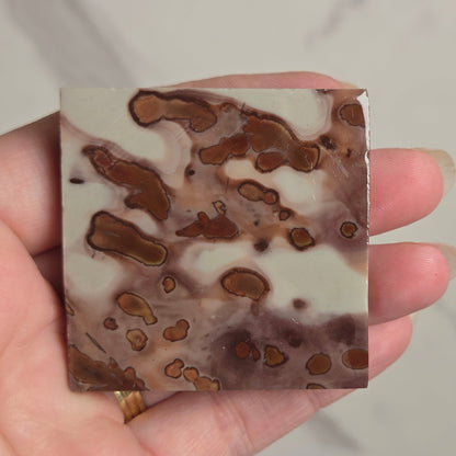 A hand holding a square decorative tile or coaster with a marbled pattern of brown, beige, and white, set against a light marble background.