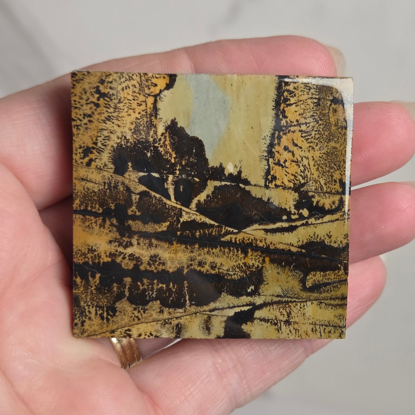 A hand holding a small square card or print featuring a landscape artwork in black, gold, and beige tones, with a light marble-like background behind it.