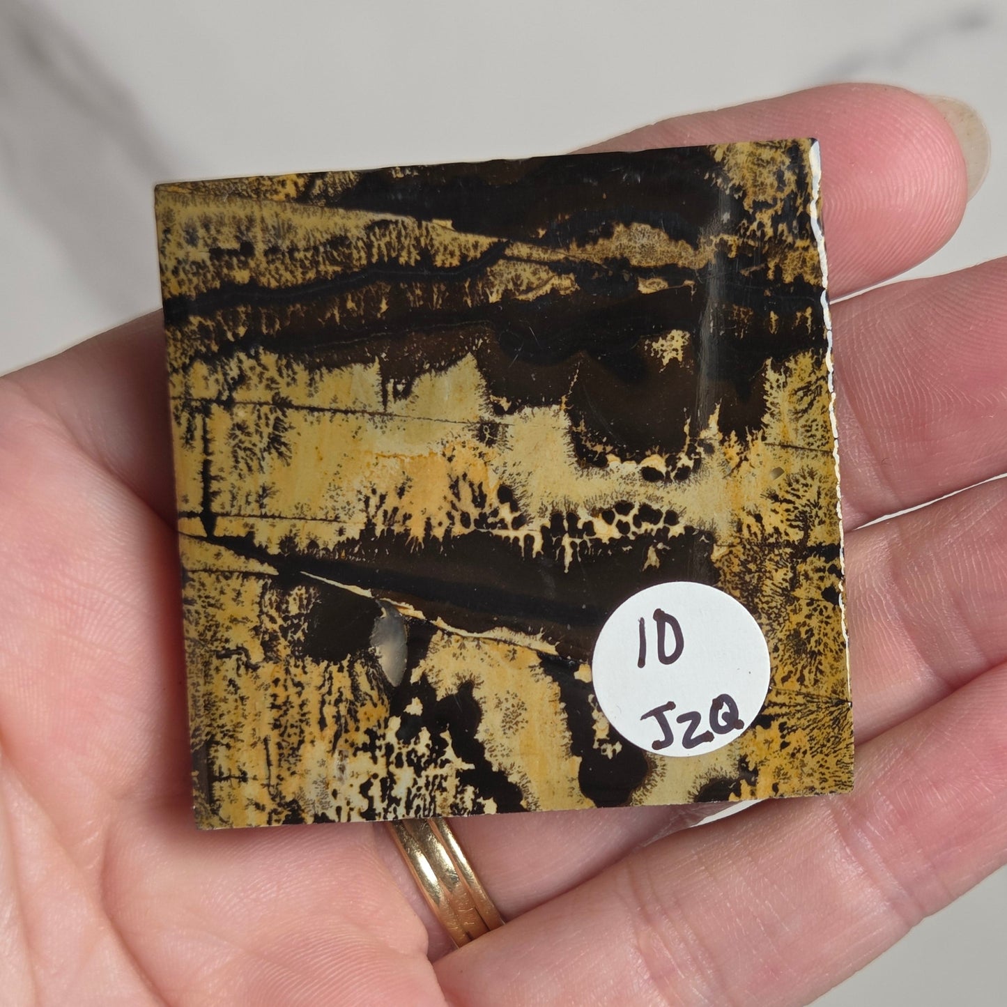 A hand holding a small square abstract art print or tile featuring a textured pattern of black, gold, and brown. A small white circular sticker with handwritten text is attached to the bottom right of the piece.