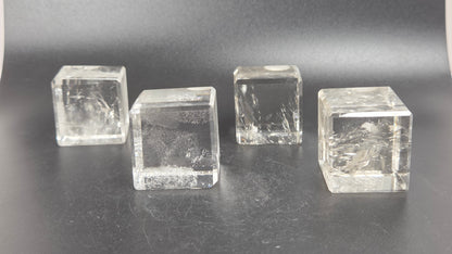 Four clear transparent cube-shaped crystal blocks with internal inclusions, arranged on a dark gray surface against a dark gray background.