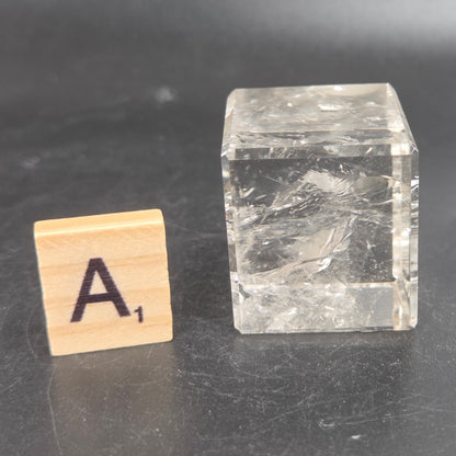 A clear, faceted clear quartz cube with visible internal textures and scratches, displayed next to a small wooden Scrabble tile with the letter 'A' on a dark gray surface.