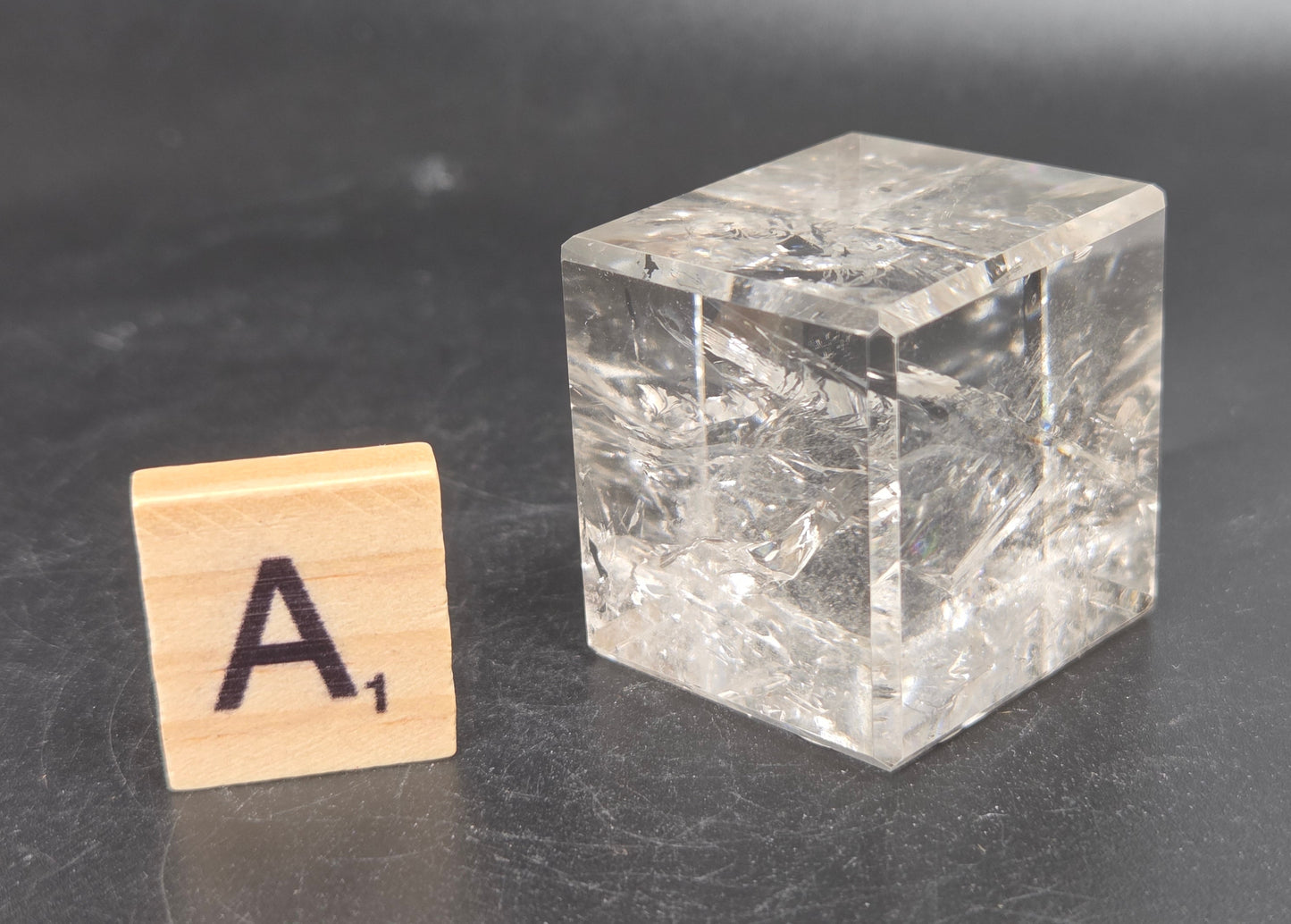 A clear, crystalline cube with visible internal texture and inclusions, placed on a dark gray surface next to a small wooden Scrabble tile showing the letter A.