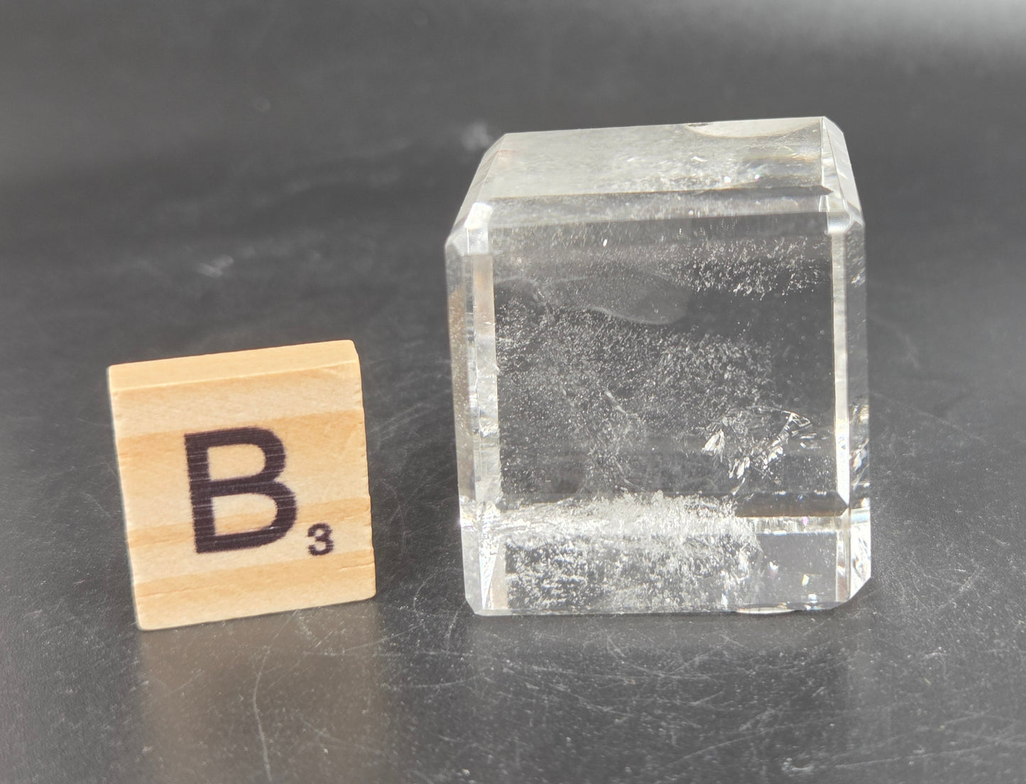 A clear rectangular glass block with a rough, textured interior, shown next to a wooden Scrabble tile marked 'B3' on a dark gray surface.