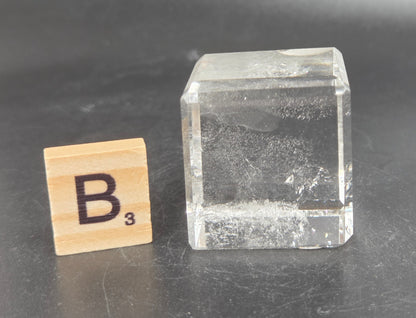 A clear rectangular glass block with a rough, textured interior, shown next to a wooden Scrabble tile marked 'B3' on a dark gray surface.