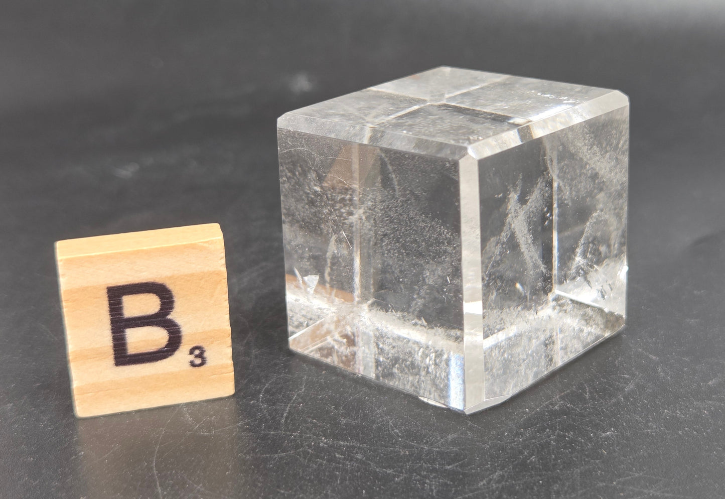 A small clear glass cube with visible internal scratches and reflections, placed on a dark gray surface next to a wooden Scrabble tile showing the letter B.