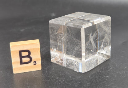 A small clear glass cube with visible internal scratches and reflections, placed on a dark gray surface next to a wooden Scrabble tile showing the letter B.