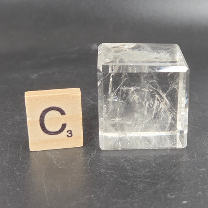 A clear rectangular crystal or glass block with visible internal inclusions, shown next to a small wooden Scrabble tile marked C3 on a dark gray surface.